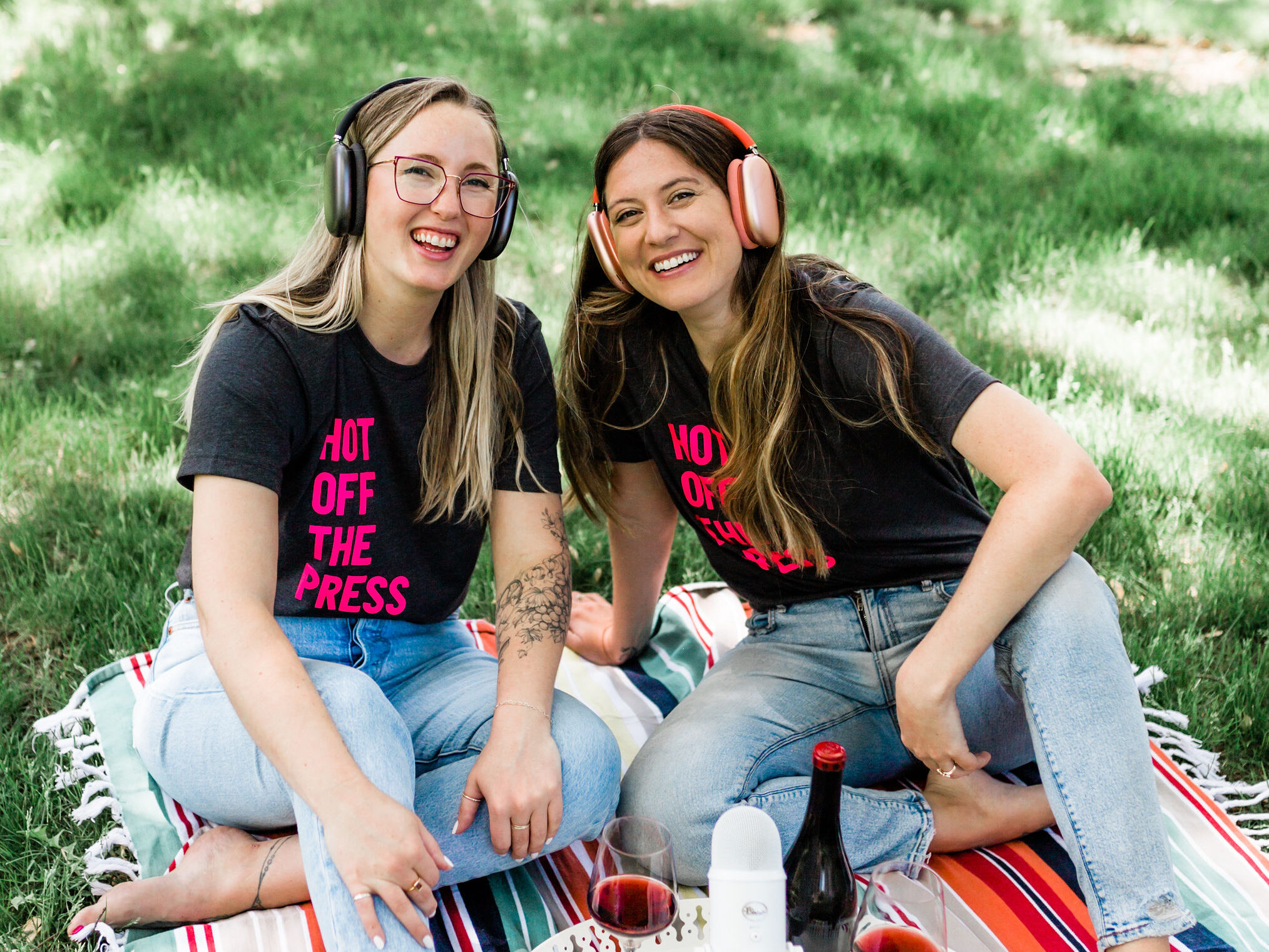The friendly faces of your podcast hosts, Moriah and Jillian, sitting outside on a picnic blanket with their podcasting gear and their Hot Off the Press branded shirts. 